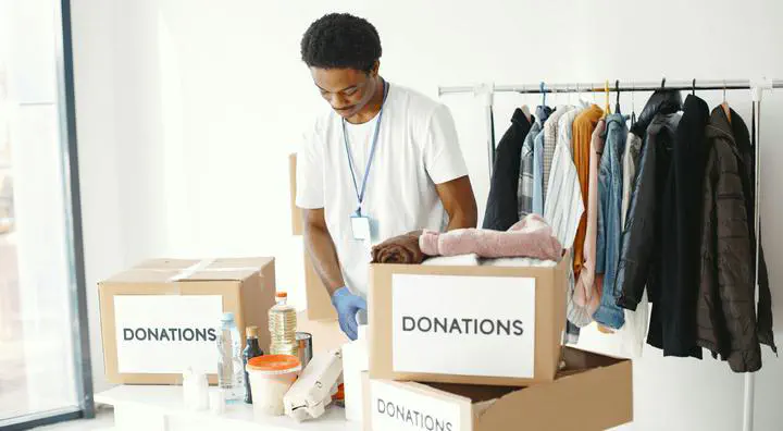 a photo of a man wearing a white t shirt standing in a room with three boxes of donations on a table and a clothing rack behind him